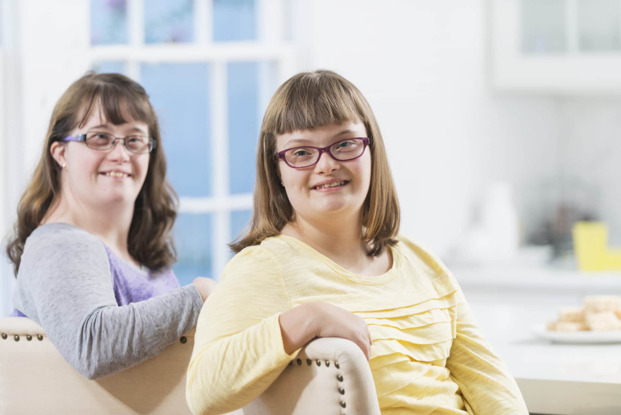 Two sisters with down syndrome sitting on chairs at home at the kitchen counter. The focus is on the younger girl, a teenager, wearing a yellow shirt.